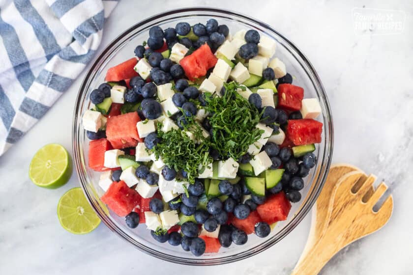 Glass bowl with watermelon, blueberries, feta, and cucumber with lime juice, cut basil and mint leaves on top.