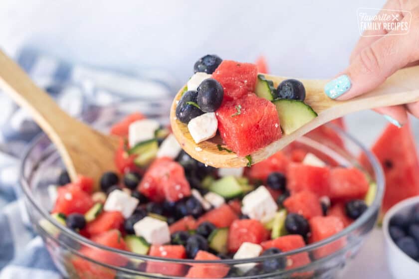 Holding a wooden spoon full of watermelon feta salad with watermelon, blueberries, feta cheese, cucumbers, milt, basil, salt and lime juice.