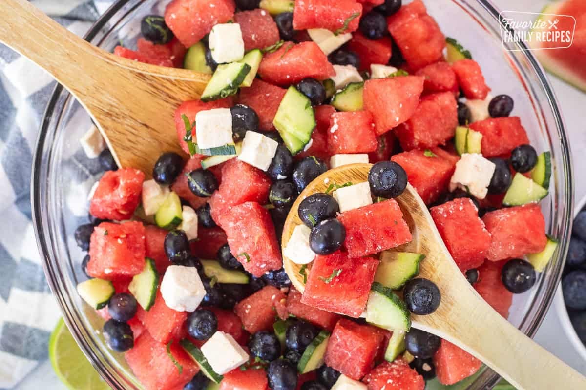 Holding a wooden spoon full of watermelon feta salad with watermelon, blueberries, feta cheese, cucumbers, milt, basil, salt and lime juice.