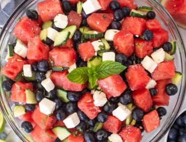 Watermelon feta salad in a glass bowl with watermelon, blueberries, feta cheese, cucumbers, milt, basil, salt and lime juice. Garnished with mint leaves.