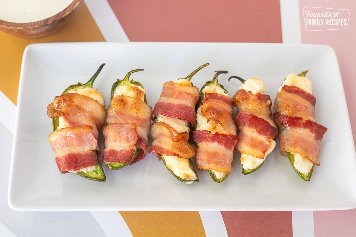 Six bacon wrapped jalapeño poppers on a plate with a bowl of Ranch dressing on the side.