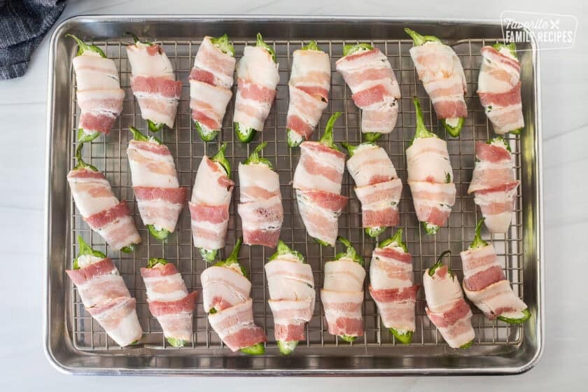 Bacon wrapped around filled jalapeños with cheese mixture on a sheet pan with wire rack.