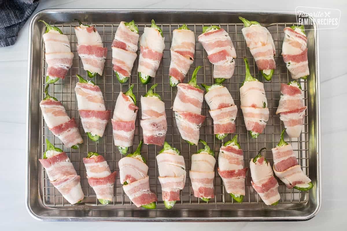 Bacon wrapped around filled jalapeños with cheese mixture on a sheet pan with wire rack.