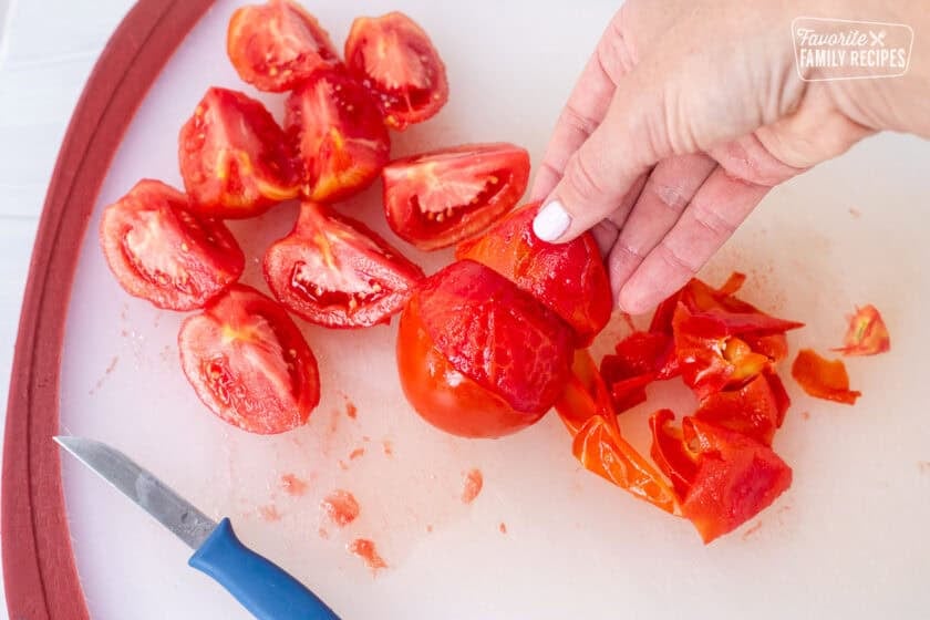 Peeling the skin off a tomato by hand on a cutting board. Quartered tomatoes on the board with a small knife.