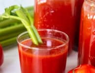 Glass of canned tomato juice with a celery stalk next to jars of tomato juice.