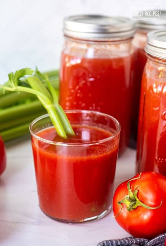 Glass of canned tomato juice with a celery stalk next to jars of tomato juice.