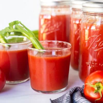 Jars of canned tomato juice with fresh tomatoes and glass of tomato juice with celery stick.