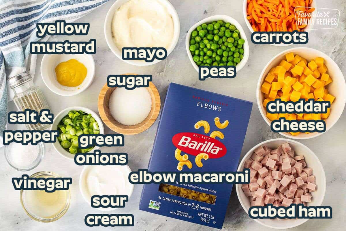 Ingredients to make Macaroni Salad including pasta, carrots, peas, cheese, ham, salt, sour cream, mayo, mustard, pepper, green onion, apple cider vinegar, pepper and sugar.