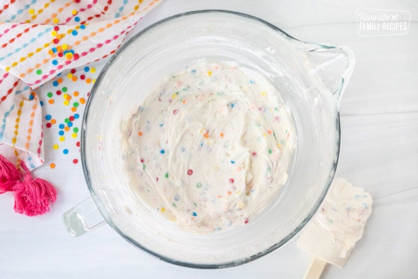 Glass mixing bowl with frosting dip mixed with sprinkles and spatula on the side.