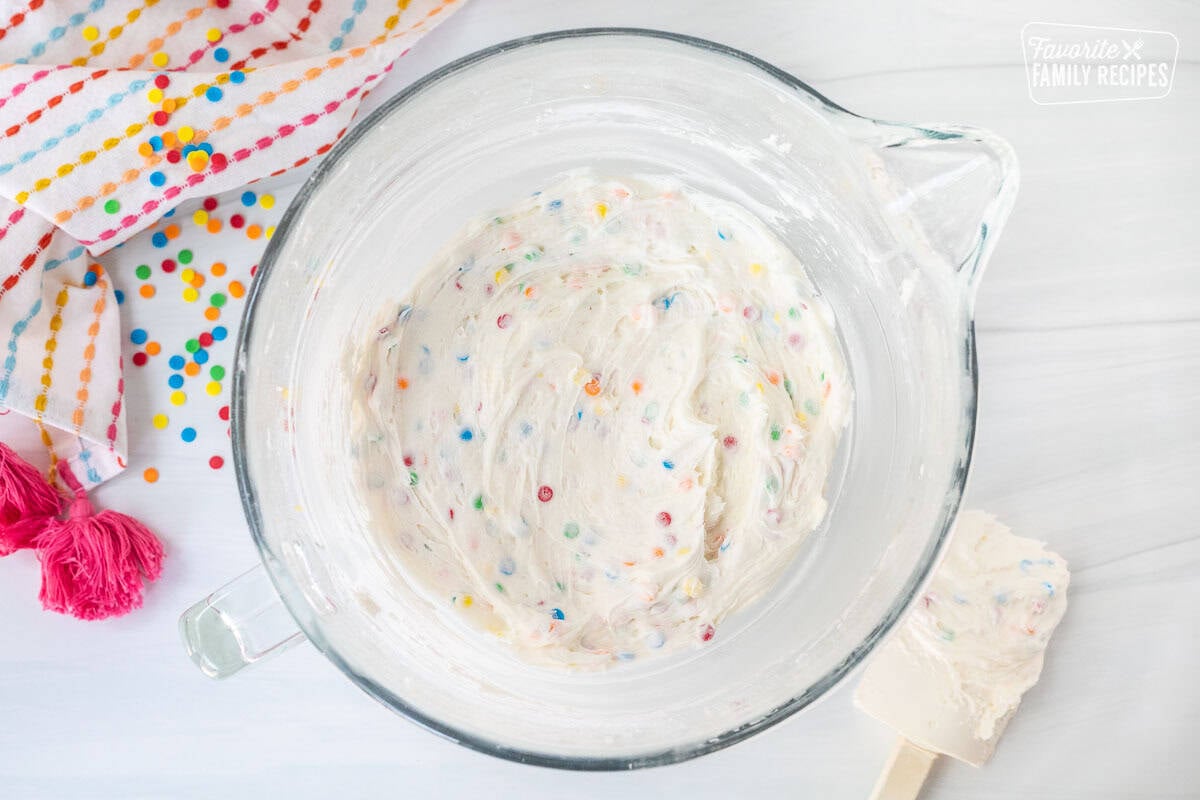 Glass mixing bowl with frosting dip mixed with sprinkles and spatula on the side.