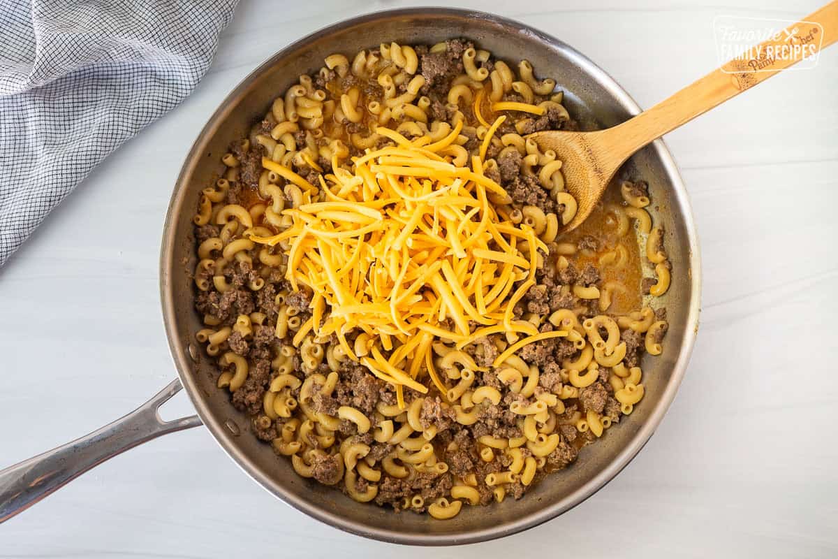 Adding shredded cheddar cheese to cooked homemade hamburger helper in a skillet.