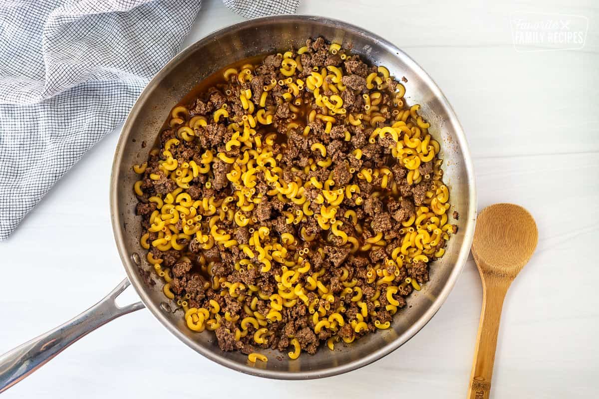Skillet with brown ground beef, beef broth, and elbow macaroni pasta stir together.