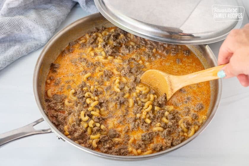 Stirring together homemade hamburger helper ingredients in a skillet with a lid on the side.