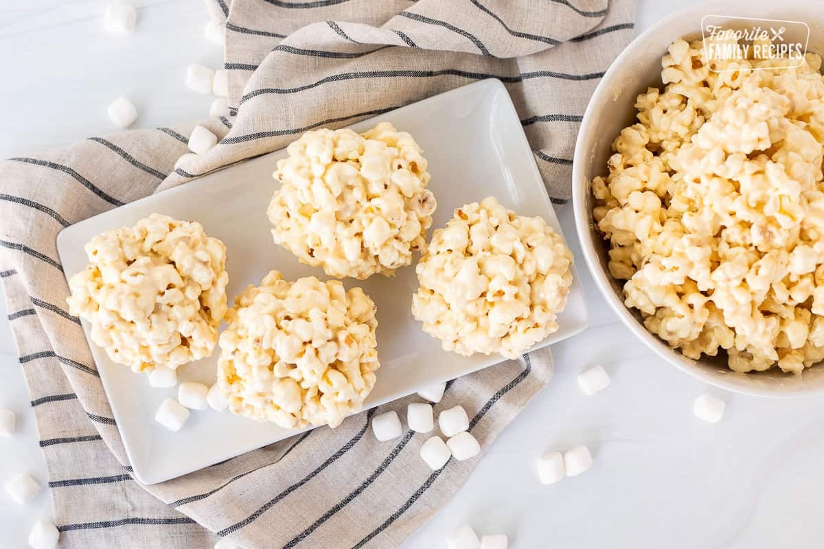 Marshmallow popcorn balls on a plate next to a bowl of marshmallow popcorn.