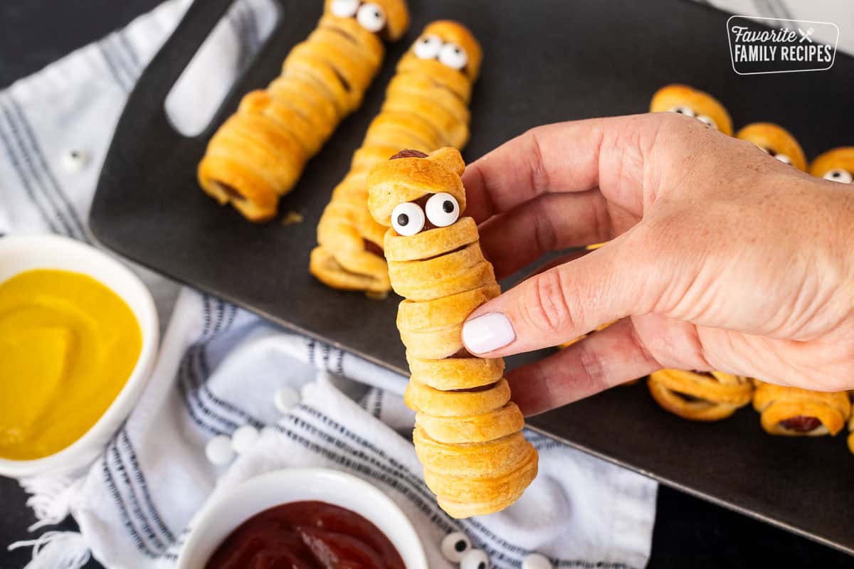 Hand holding a mummy hot dog with sprinkle eyeballs. Ketchup and mustard on the side.