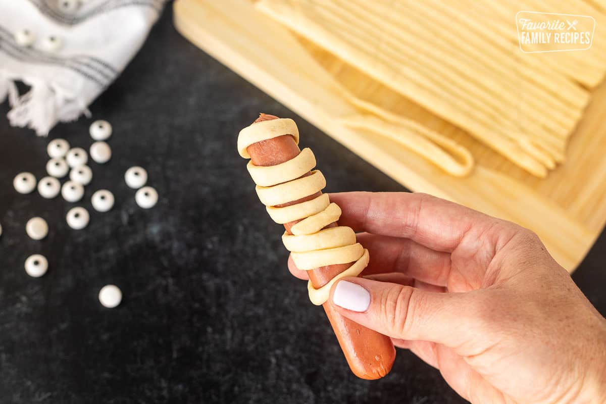 Wrapping a hot dog with strips of crescent roll strips.