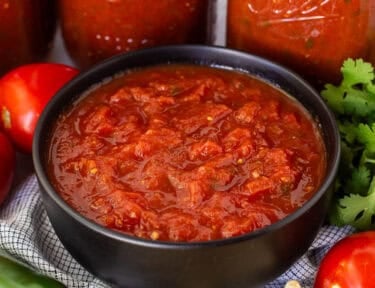 Bowl of savory canned salsa next to jars of salsa and chips on the side.