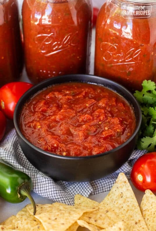 Bowl of savory canned salsa next to jars of salsa and chips on the side.