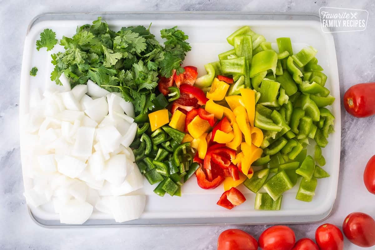 Cutting board with chopped green bell peppers, yellow peppers, red peppers, jalapeños, cilantro, white onion, and Serano peppers.