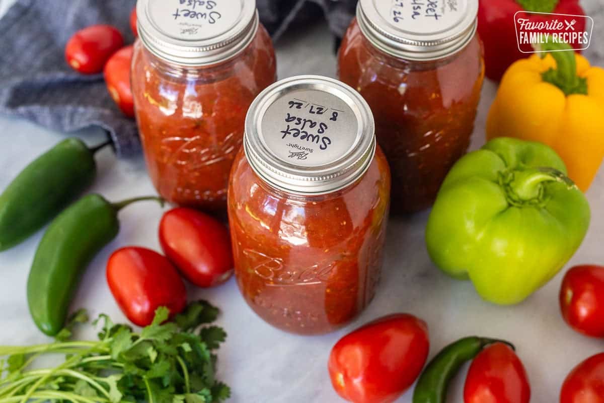Labeled jars of sweet canned salsa surrounded by fresh vegetables.