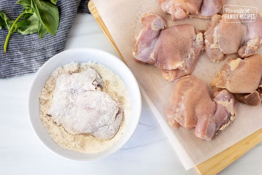 Chicken thigh covered in flour in a bowl next to chicken thighs on a cutting board seasoned with salt and pepper.
