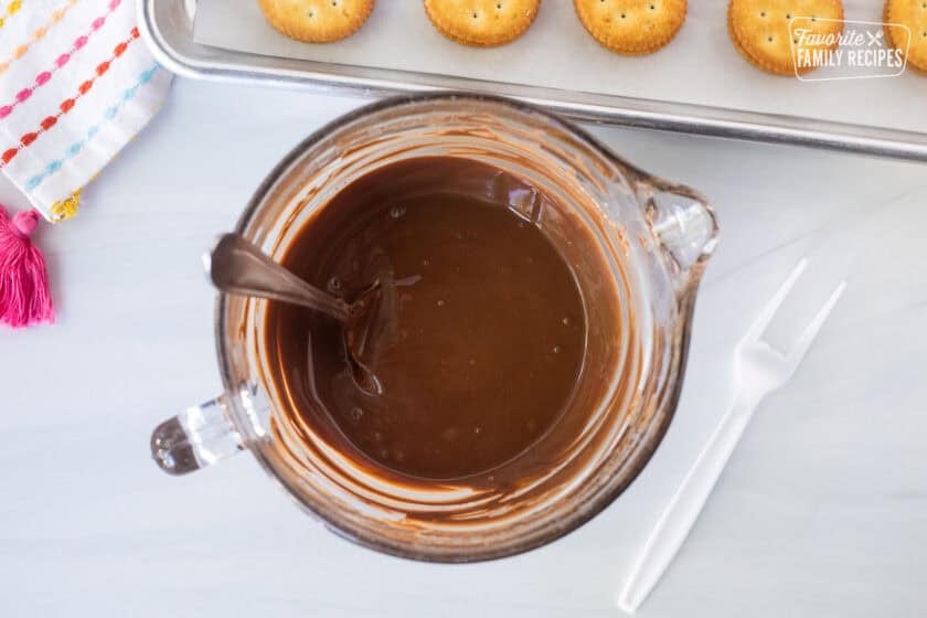 Glass bowl with melted milk chocolate and plastic broken off fork on the side. Ritz crackers sandwiched on the side.