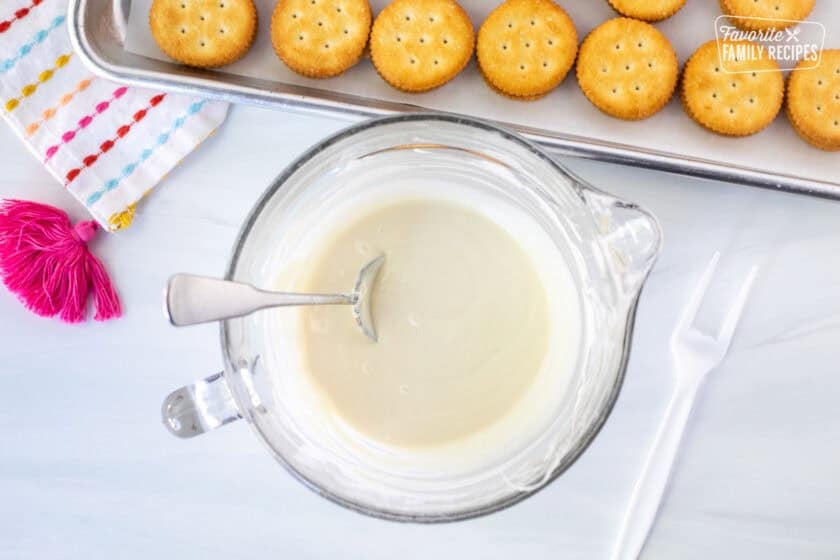 Glass bowl with melted white chocolate and plastic broken off fork on the side. Ritz crackers sandwiched on the side.