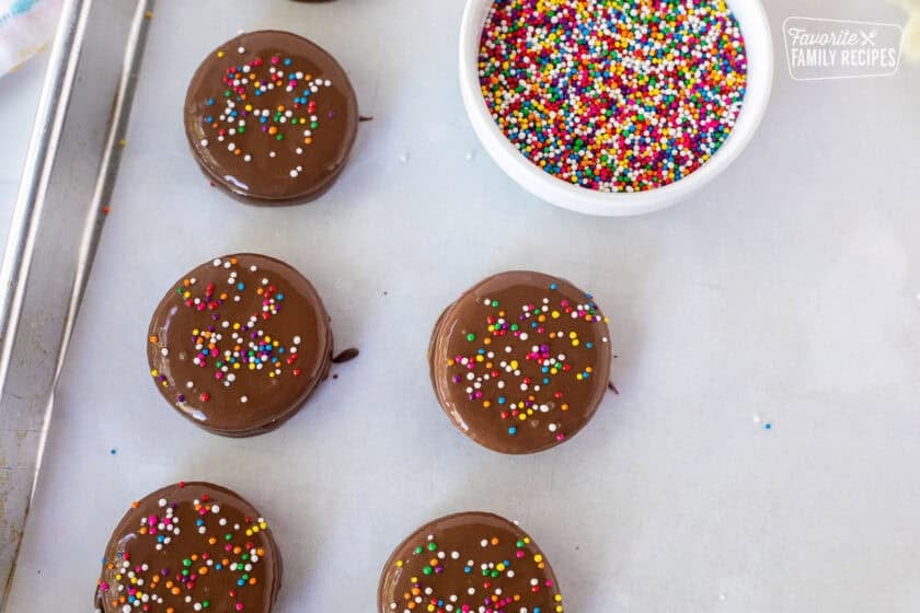 Milk chocolate covered Ritz crackers with sprinkles on top next to a bowl of sprinkles drying on parchment paper.