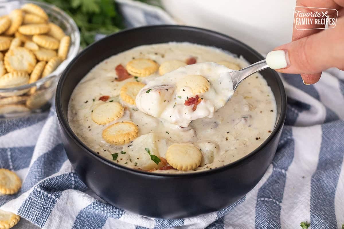 Spoon lifting out of a bowl of clam chowder with fresh clams and garnished with bacon and crackers.