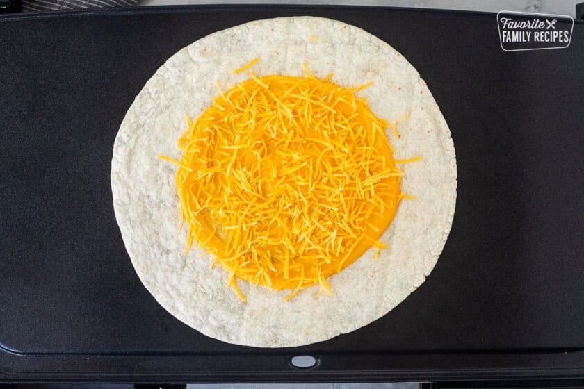 Skillet with tortilla topped with nacho cheese and shredded cheddar cheese on top.
