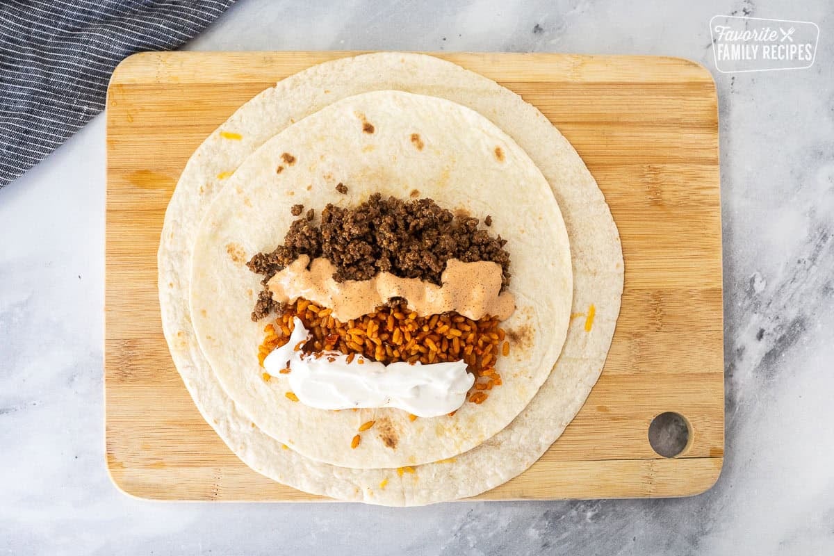 Placing ground beef, rice, sour cream and chipotle sauce inside the smaller tortilla for a quesarito.