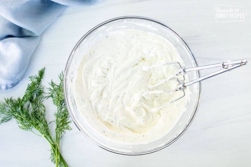 Glass mixing bowl with cream cheese, Greek yogurt, fresh dill and pickle juice mixed with electric beaters.