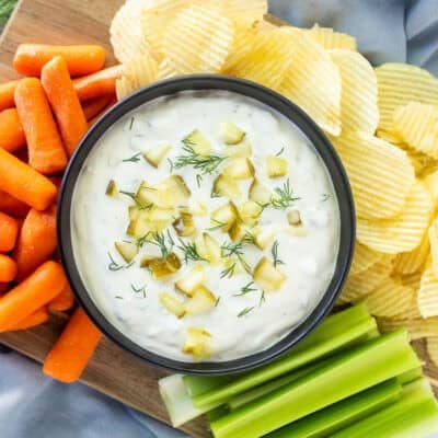 Dill pickle dip in a bowl surrounded by chips, celery sticks and baby carrots.