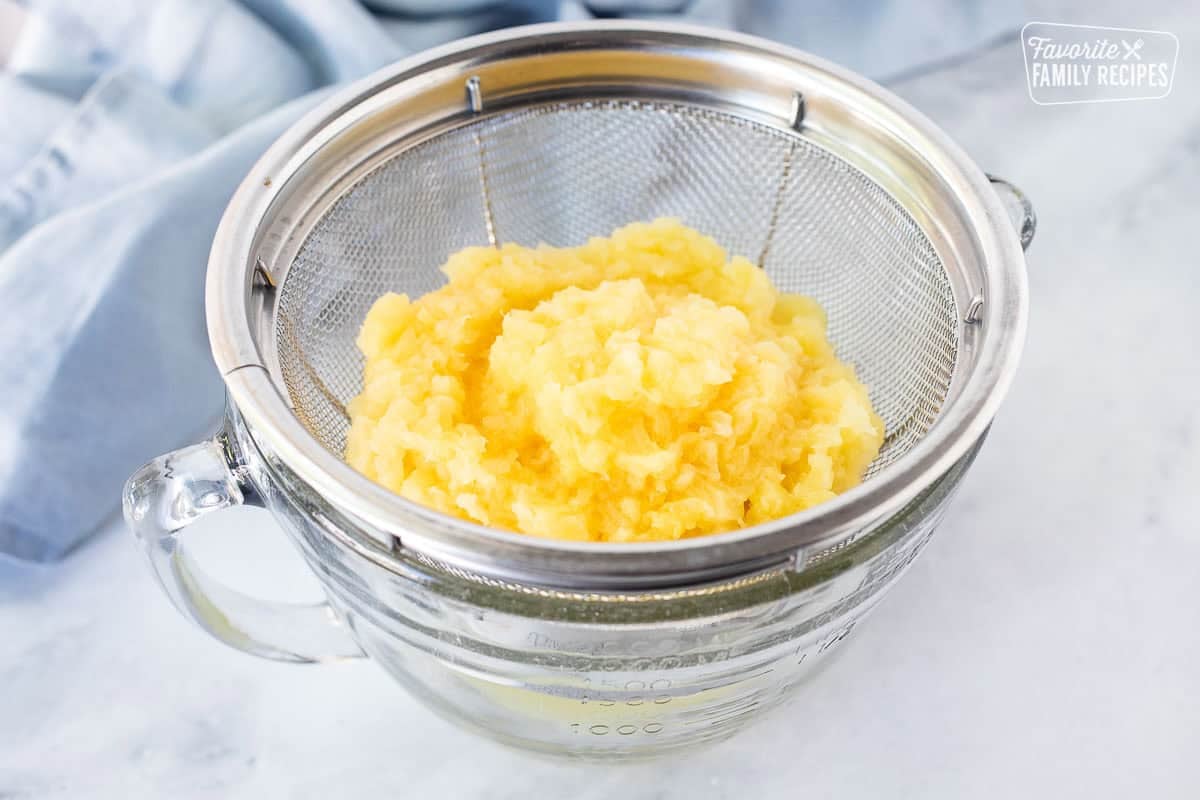 Crushed pineapple draining through a colander into a bowl.