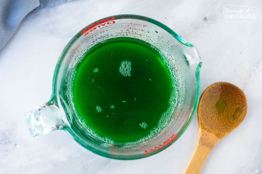 Bowl of lime jello mixed with hot water and wooden spoon on the side.
