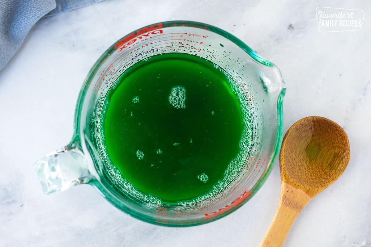 Bowl of lime jello mixed with hot water and wooden spoon on the side.