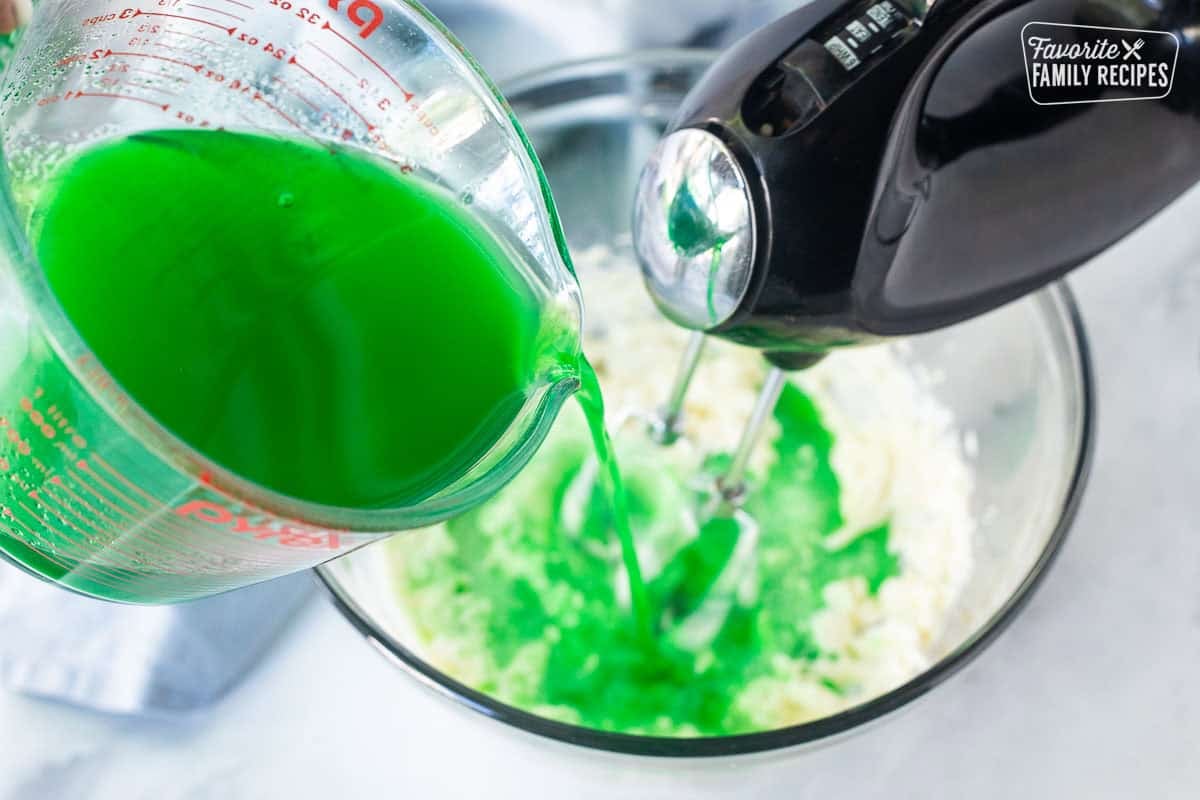 Pouring lime jello into bowl of cream cheese mixture while whipping with electric beaters.