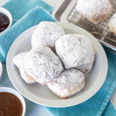 Plate of New Orleans style beignets covered in powdered sugar with three small bowls of dipping sauces including caramel sauce, chocolate sauce, and red raspberry preserves.