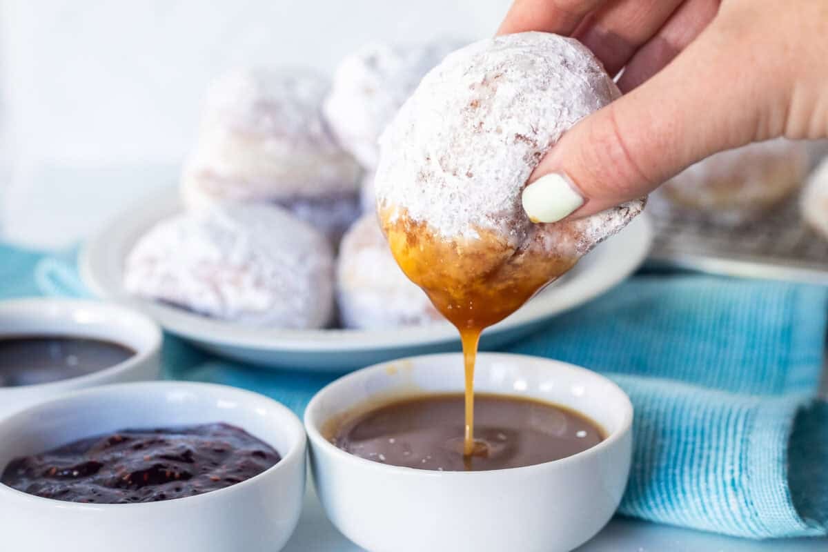 New Orleans beignet covered in powdered sugar held in hand and dipped into a bowl of caramel sauce.