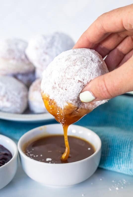 New Orleans beignet covered in powdered sugar held in hand and dipped into a bowl of caramel sauce.