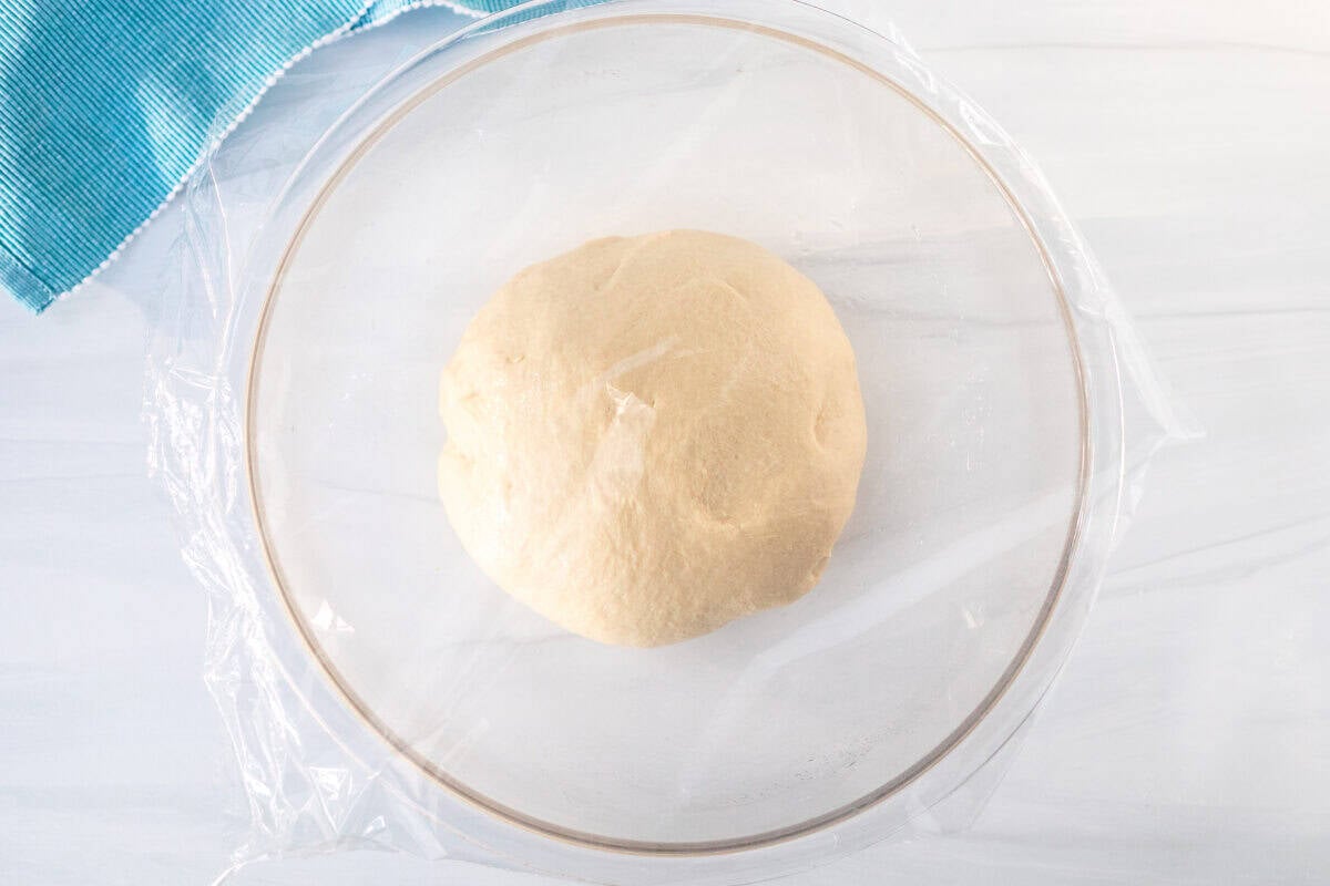 Large glass bowl with a ball of dough and covered with plastic wrap.