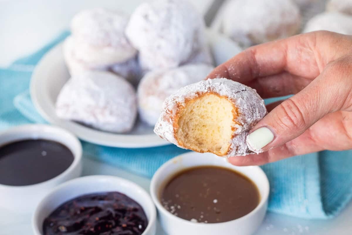 Hand holding a New Orleans style beignet missing a bite. Three bowls of dipping sauces including caramel, red raspberry preserves and chocolate sauce.
