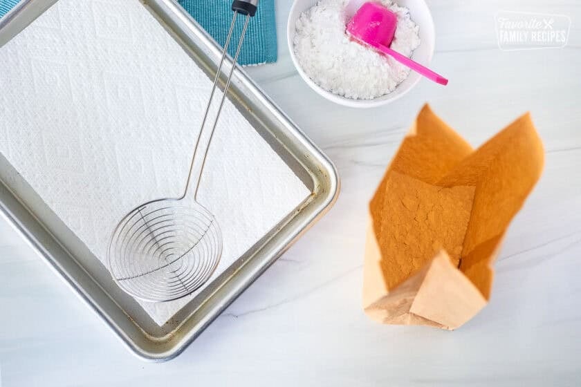 Sheet pan lined with paper towels, strainer, bowl of powdered sugar with scoop and paper bag with powdered sugar inside.