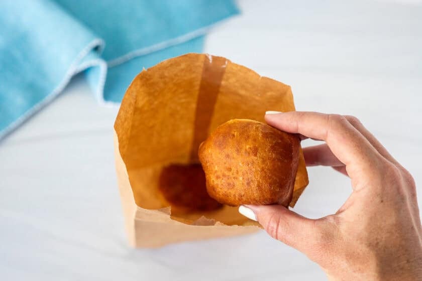 Placing a fried New Orleans style beignet into a paper bag with powdered sugar in inside.