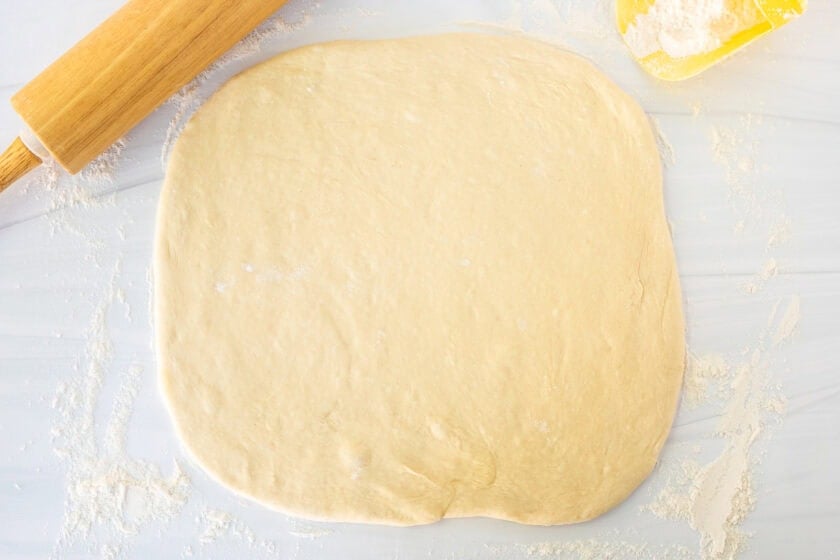 Square piece of beignet dough rolled out on a floured surface with a rolling pin on the side.