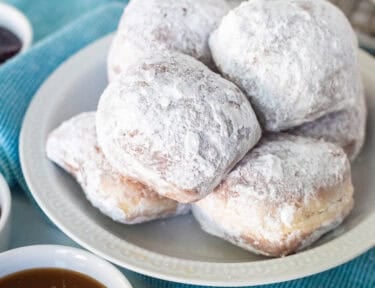 Plate of New Orleans style beignets covered in powdered sugar with three small bowls of dipping sauces including caramel sauce, chocolate sauce, and red raspberry preserves.