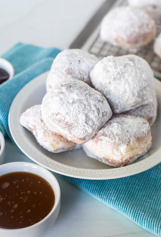 Plate of New Orleans style beignets covered in powdered sugar with three small bowls of dipping sauces including caramel sauce, chocolate sauce, and red raspberry preserves.