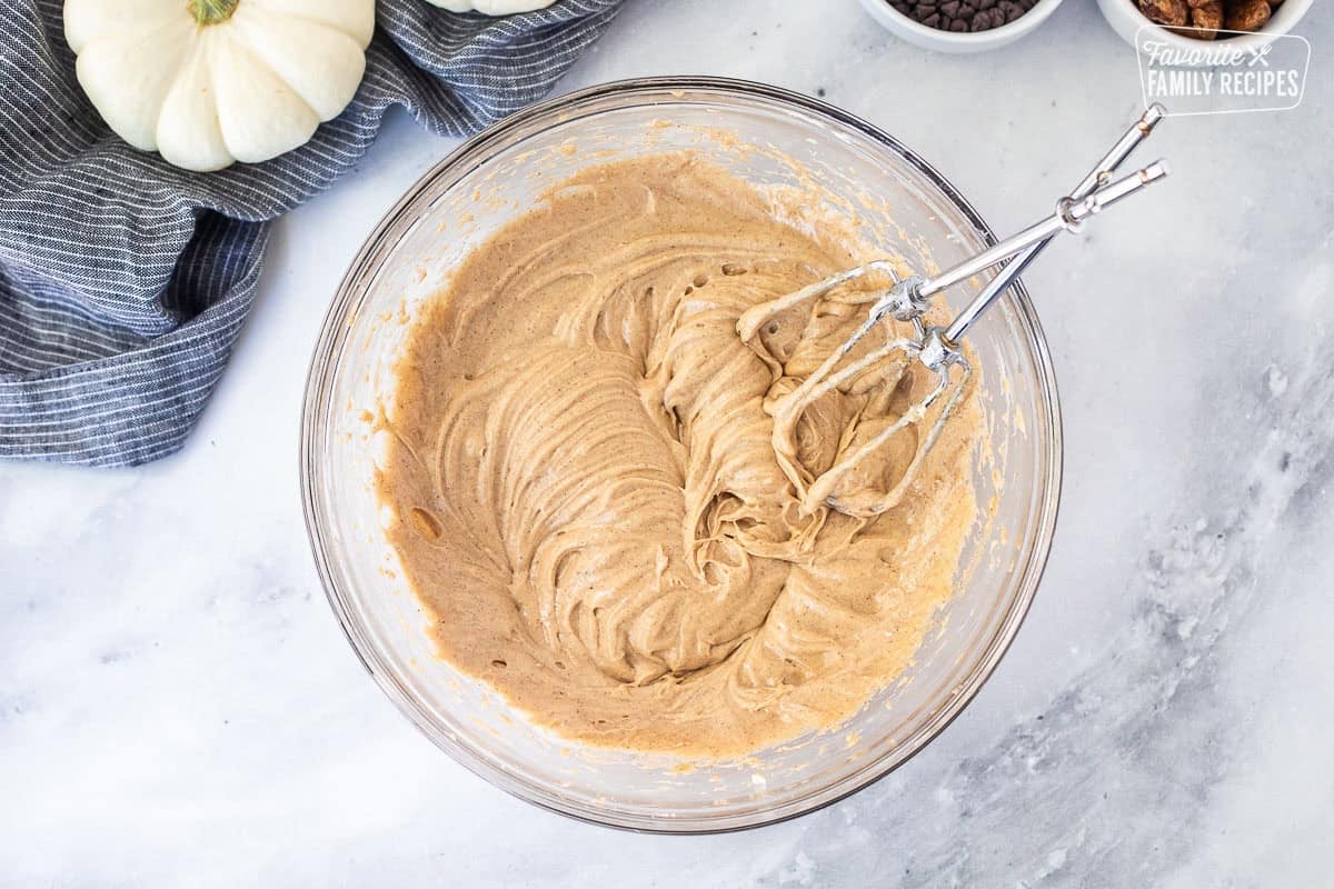 Glass mixing bowl of whipped cream cheese and pumpkin pie spice with electric beaters.