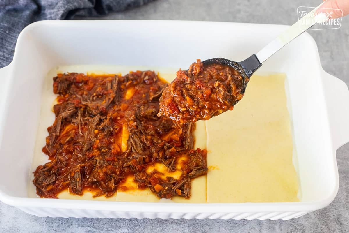 Spooning short rib ragu sauce over lasagna noodles in a baking dish.