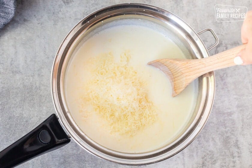 Adding shredded cheeses to béchamel sauce in a saucepan with a wooden spoon.